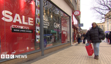 A shopper passes a shop window with red sale, 70% off signs on a high street. There are other people walking in the background, some with shopping bags and a Halifax sign. There are tree branches on the right of the picture.