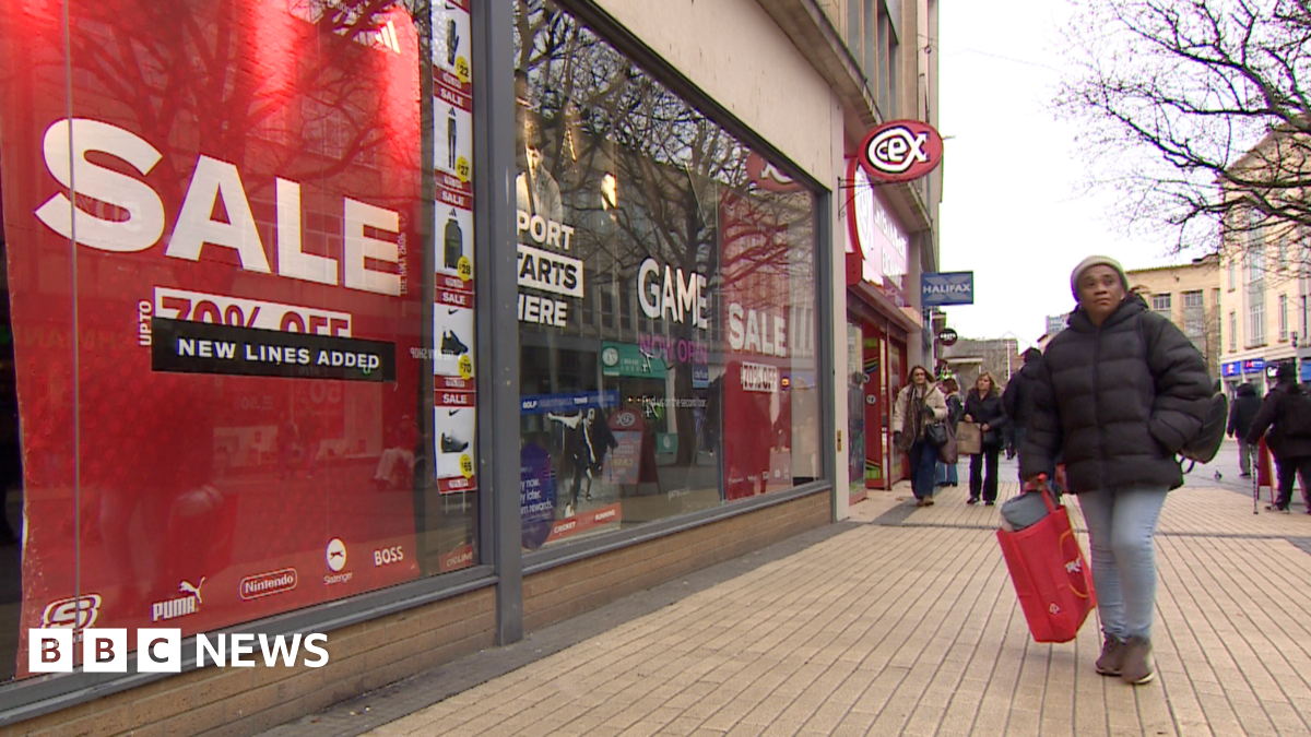 A shopper passes a shop window with red sale, 70% off signs on a high street. There are other people walking in the background, some with shopping bags and a Halifax sign. There are tree branches on the right of the picture.
