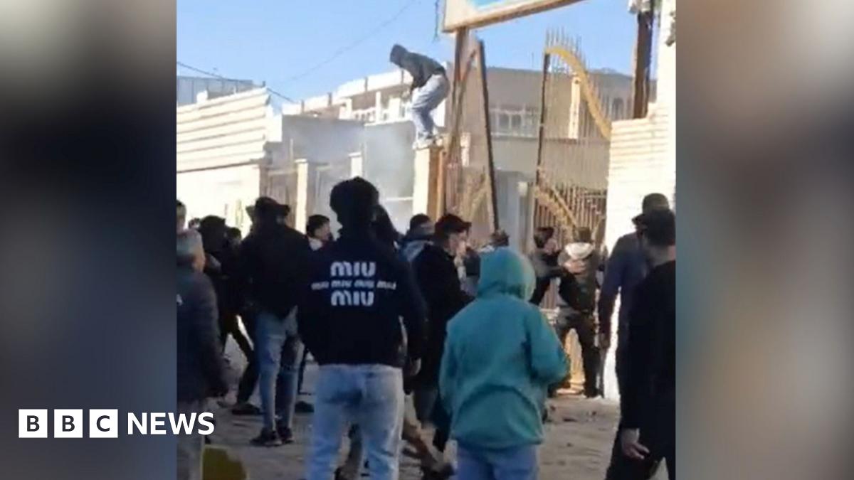 In a screengrab taken from footage, protesters facing away from the camera are gathered outside a building with large yellow gates which are leaning at a wonky angle. One hooded protester appears to be jumping off the gates.