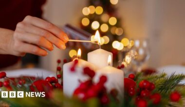 A person lights a white candle with a match on a festive table decorated with holly and red berries, with a softly lit Christmas tree blurred in the background.