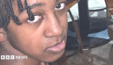A snapshot of a young man with braided hair, looking into the camera from below. He appears to be in a bar or cafe, with the bases of tables visible in the background.