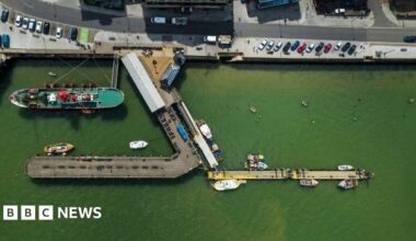 An overhead view of the Ha'penny Pier, Harwich, which juts out into the port.  There are a number of boats on green looking water and cars parked along the quay.