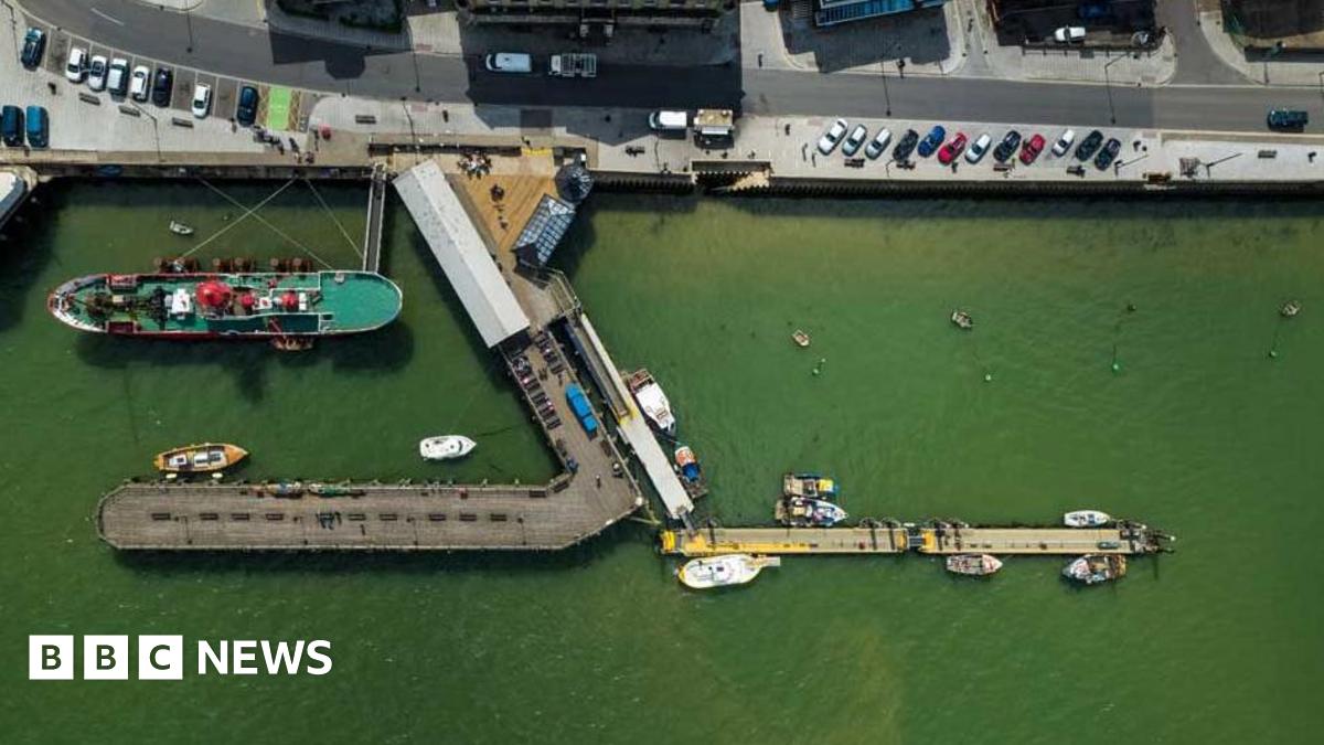 An overhead view of the Ha'penny Pier, Harwich, which juts out into the port.  There are a number of boats on green looking water and cars parked along the quay.