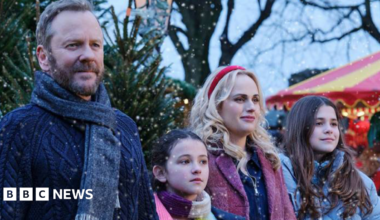 The production still shows 4 actors dressed in winter clothes with Christmas trees behind them. A red and yellow coloured fairground ride is on the right of the image. It is snowing.