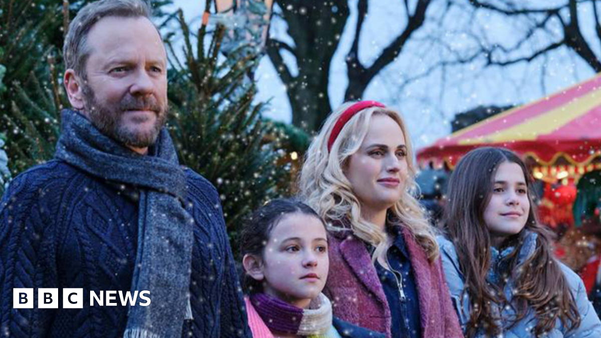 The production still shows 4 actors dressed in winter clothes with Christmas trees behind them. A red and yellow coloured fairground ride is on the right of the image. It is snowing.