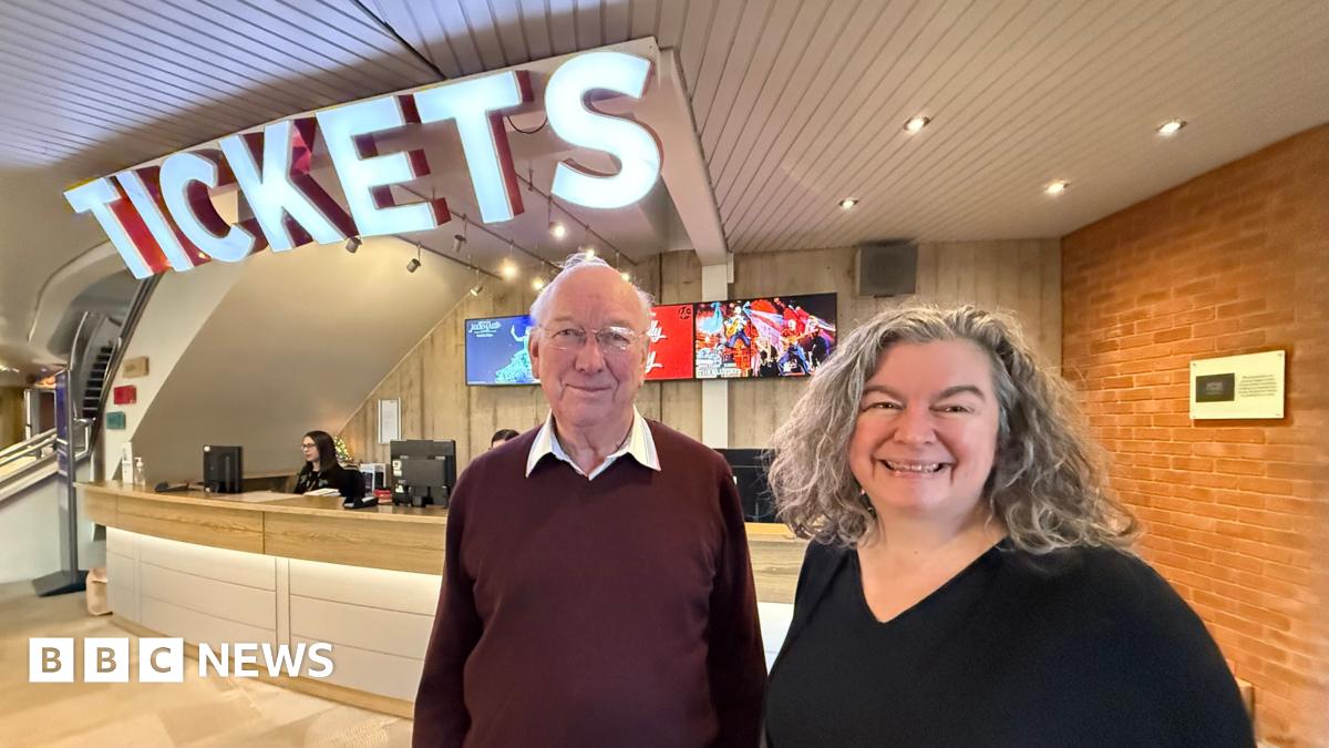 John Sambrook, a man wearing glasses and a maroon coloured jumper. He is standing next to a brick-building with white lettering on the side which reads New Vic Theatre.