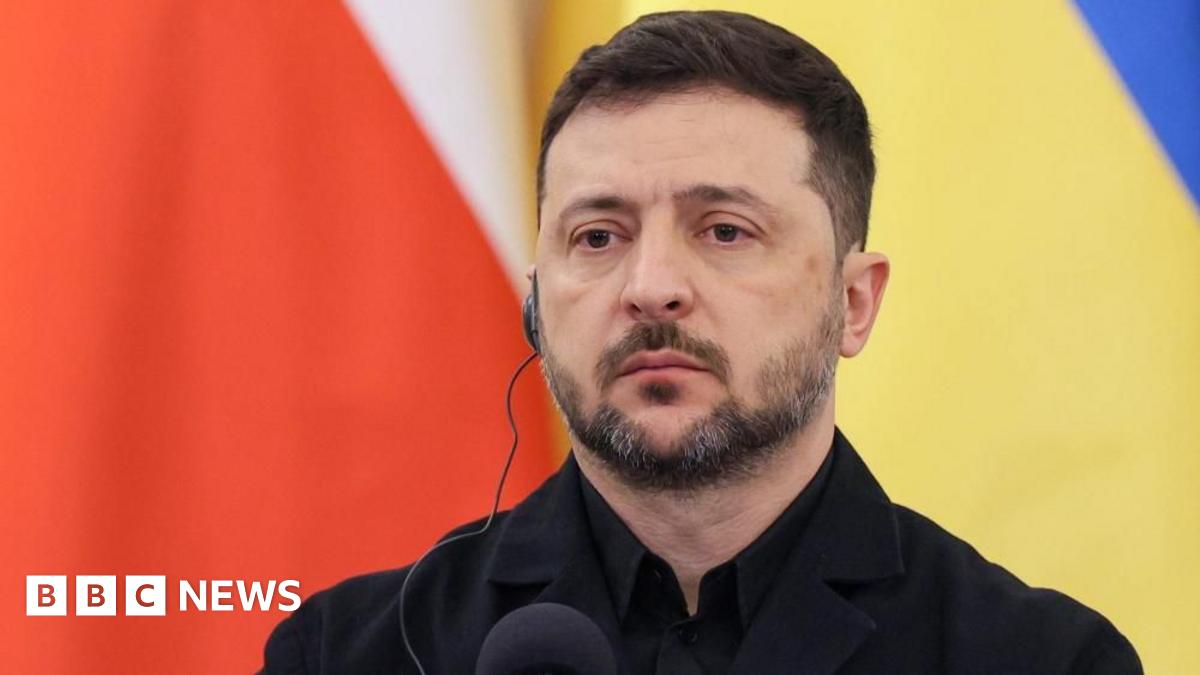 Zelensky, who has a serious expression, wears an earpiece and a black jacket with a black shirt as he stands in front of a microphone at a press conference