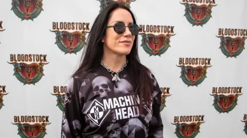Vicky Hungerford A woman with long black hair, sunglasses, a large silver necklace and a purple and black Machine Head top with skulls pictured on it. She is standing in front of a wall which has the Bloodstock Festival logo on it repeatedly.