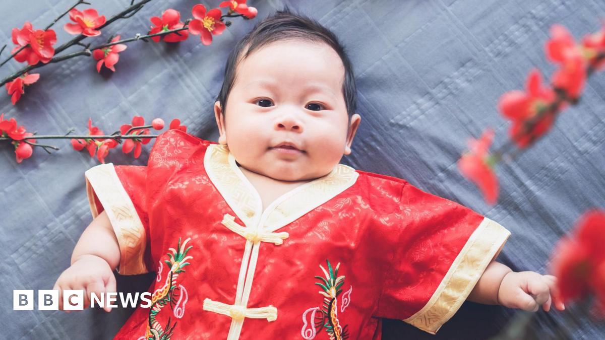 A baby lying down on a patterned grey cloth while dressed in a red traditional Chinese outfit with gold linings. Some red flowers surround him.