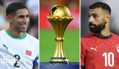 A triptych showing a vertical images of a smiling Achraf Hakimi, the Africa Cup of Nations trophy on a blue plinth and Mohamed Salah. Hakimi wears a white Morocco shirt with green trim displaying the number two on the chest, while Salah, in a red Egypt shirt bearing a number 10 on the chest, is turning his head to one side and has a serious expression on his face. Both men are visible from stomach up