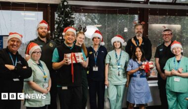 A group of hospital staff in green and blue medical uniforms are wearing read and white Santa hats and antlers.