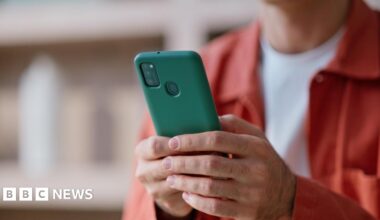 A person wearing a bright, orange jacket and white t-shirt types on a smartphone held in their hands.
