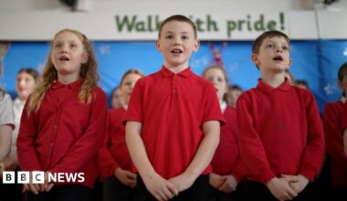 Students at Mersey Drive Community Primary School sing in their school hall. It's a close-up shot of three singing primary age children, two boys centre and right, and one girl on the left, all in red school uniform tops. More children stand behind them in rows in the choir.