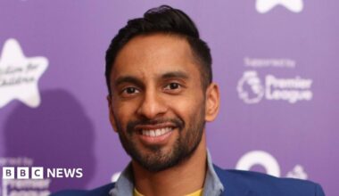 Bobby Seagull, a man with short black hair, wearing a yellow t-shirt, with a grey half-zipped jumper and a blue blazer, smiles for the camera while standing in front of a purple background.