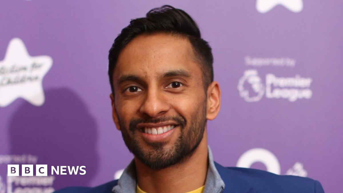 Bobby Seagull, a man with short black hair, wearing a yellow t-shirt, with a grey half-zipped jumper and a blue blazer, smiles for the camera while standing in front of a purple background.