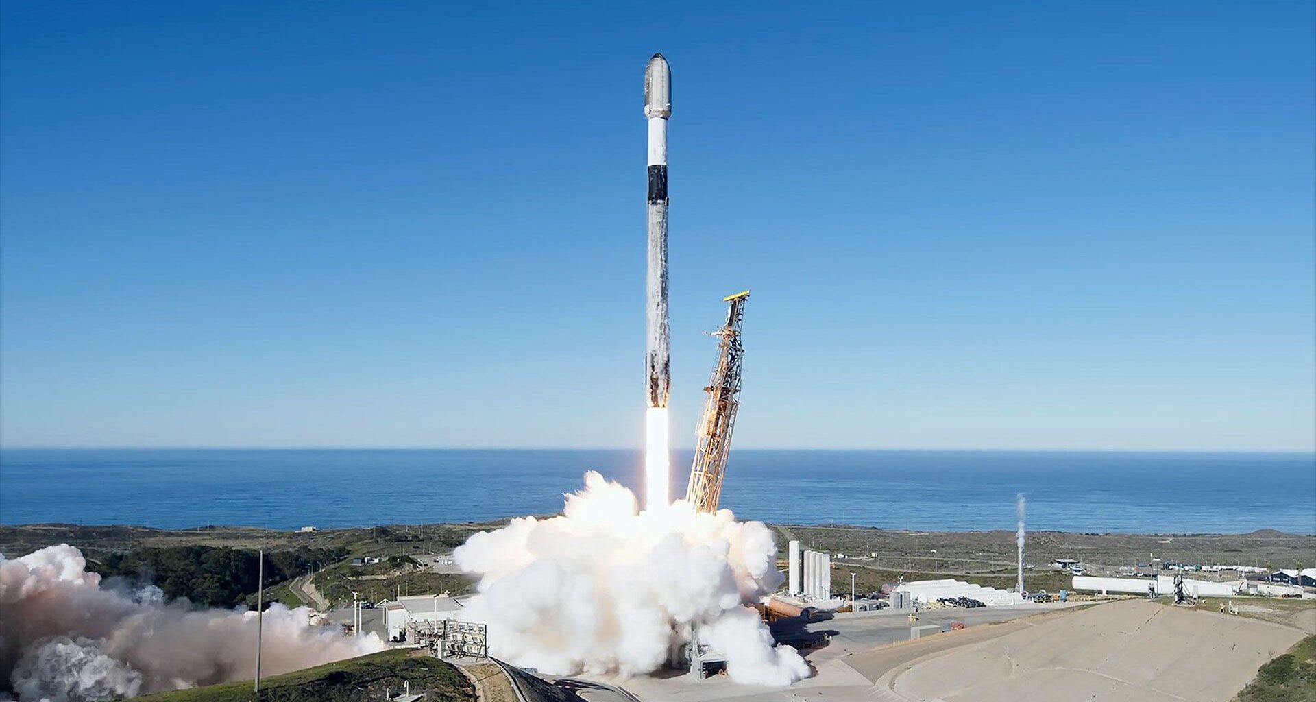 a white rocket with a black interstage lifts off from its oceanside launch pad into a clear blue sky