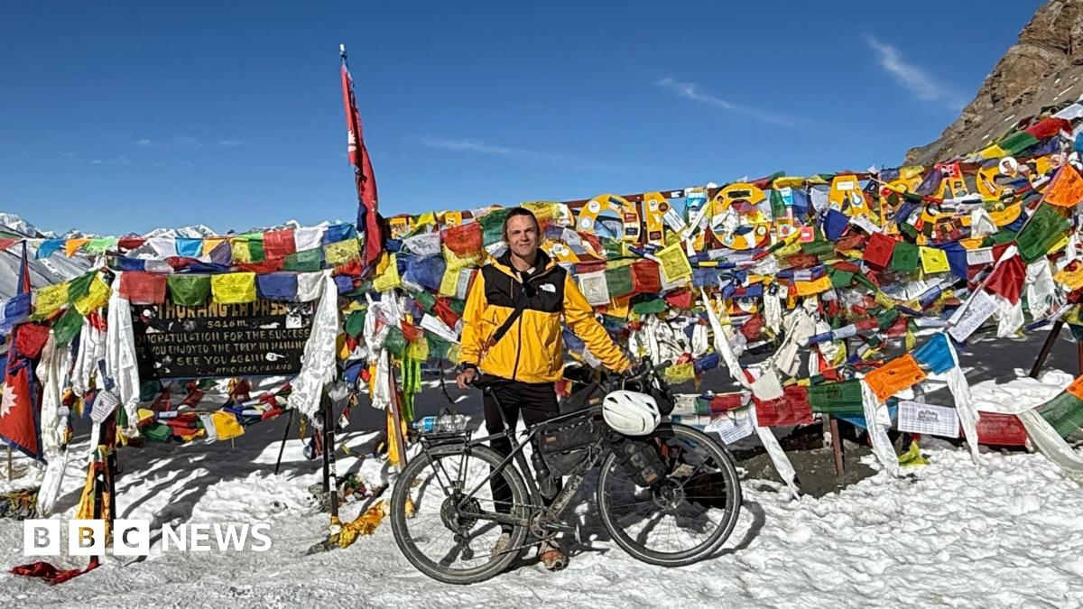 Spencer Wragg, in a yellow coat, standing over his bike on a snowy mountain surrounded by a number of colourful flags