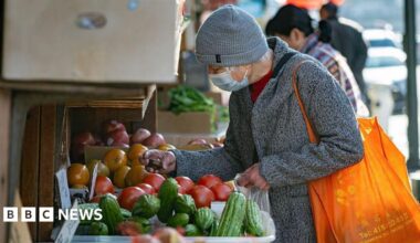 A shopper in a grey winter coat and grey hat, carrying an orange bag, browses tomatoes, cucumbers and other produce in the Chinatown neighborhood of San Francisco, California, US, on Monday, Dec. 15, 2025.