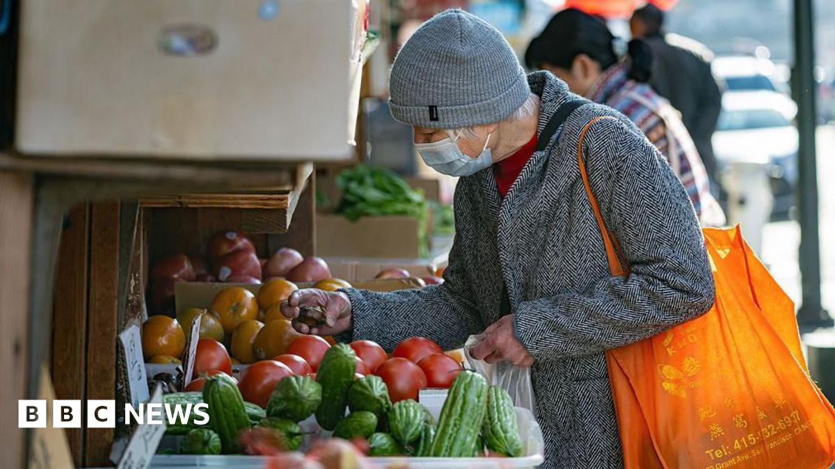 A shopper in a grey winter coat and grey hat, carrying an orange bag, browses tomatoes, cucumbers and other produce in the Chinatown neighborhood of San Francisco, California, US, on Monday, Dec. 15, 2025.