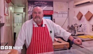 Butcher Paul Schofield stands behind his counter in The Quality Butcher on Market Street in Penistone. He is wearing a red and white striped butcher's apron. A yellow chopping board and knife are to the right of his hand, which is resting on the glass counter.