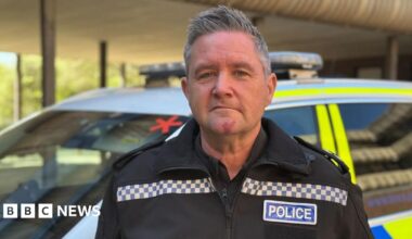 A man with short grey hair that is styled in an upward fashion is wearing a black jacket and shirt with 'POLICE' in blue and white on the side. He is pulling a serious expression into the camera and behind him is a white, yellow and blue police car parked up that is blurred.