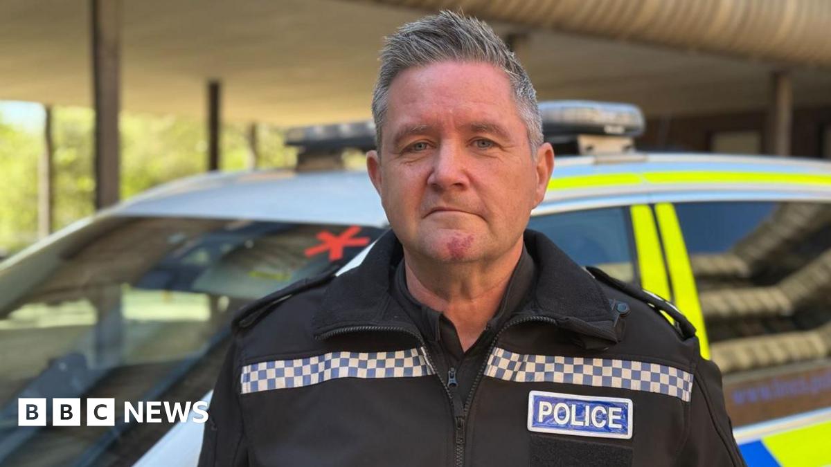 A man with short grey hair that is styled in an upward fashion is wearing a black jacket and shirt with 'POLICE' in blue and white on the side. He is pulling a serious expression into the camera and behind him is a white, yellow and blue police car parked up that is blurred.