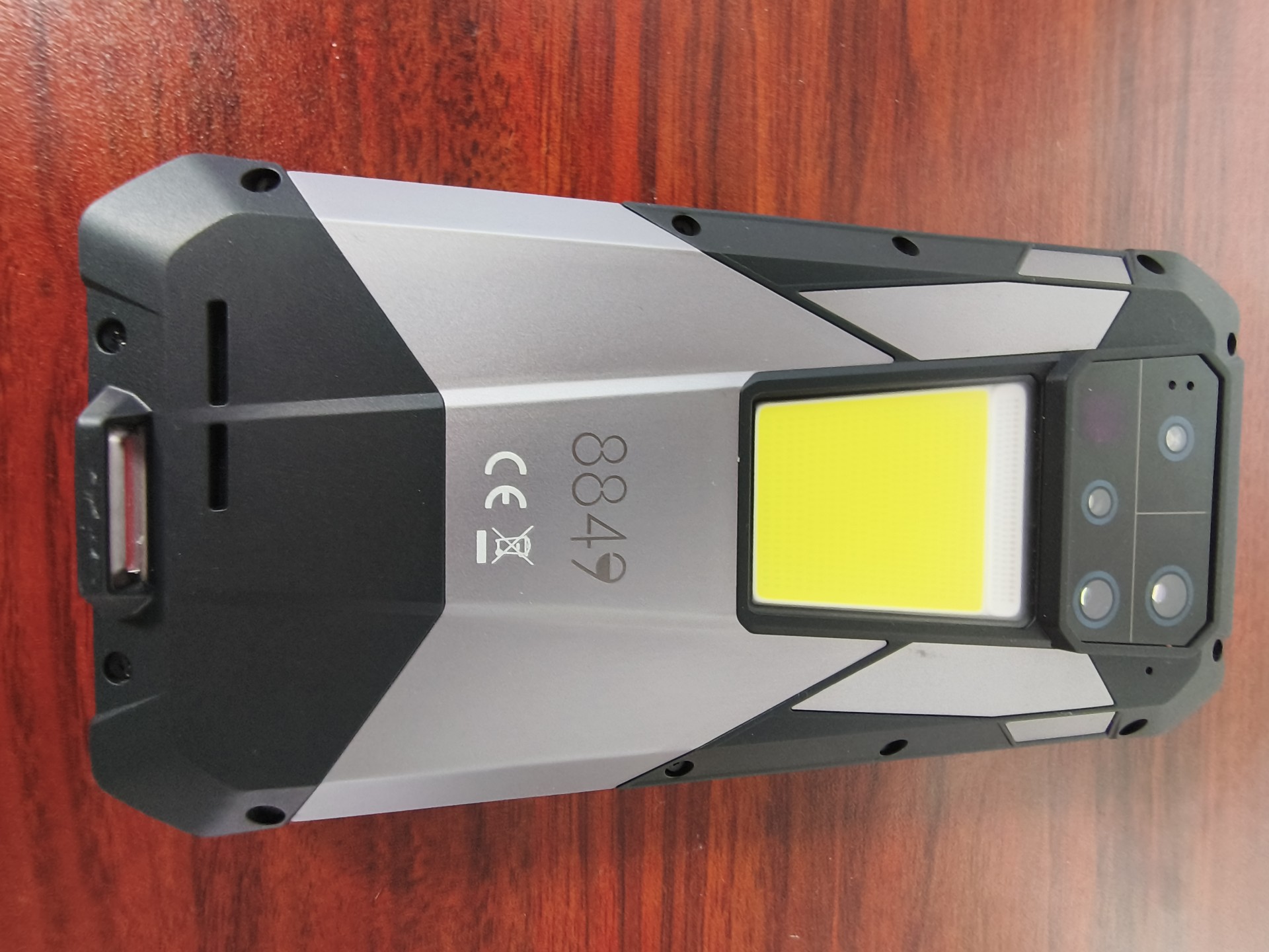 A vertical view of the back of the Tank 4 Pro rugged smartphone, featuring a large, bright yellow square COB LED light, a camera module, and a silver and black geometric panel design with a CE mark.