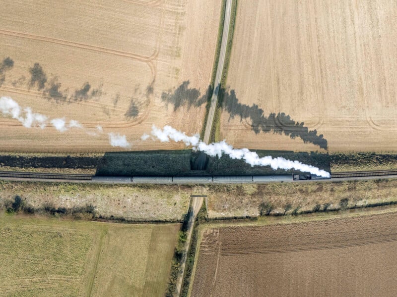 Aerial view of a steam train traveling through farmland, with white smoke trailing behind. The tracks cut straight through tan and brown fields, and a narrow road crosses over the railway.