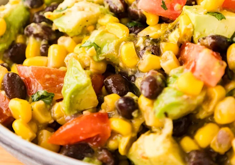 Close-up of a salad with black beans, corn, avocado, tomato, and chopped herbs mixed together in a bowl.