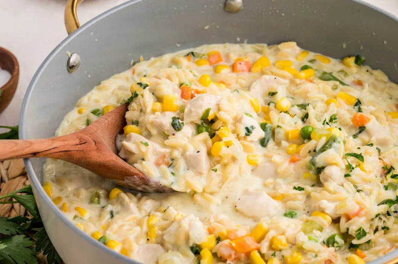 A creamy chicken and vegetable skillet dish with corn, carrots, peas, and herbs is being stirred with a wooden spoon.