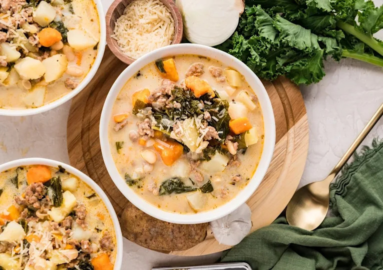 A bowl of creamy soup with sausage, potatoes, kale, carrots, and beans, garnished with shredded cheese, surrounded by fresh greens, parmesan, and a gold spoon.