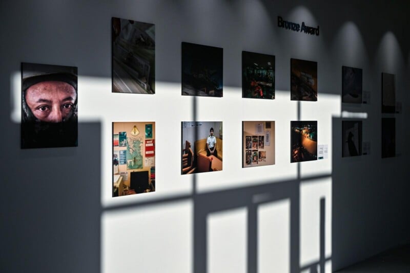 A gallery wall displays twelve photographs under the label "Bronze Award." Sunlight streams through window panes, casting sharp shadows across the images and the white wall.