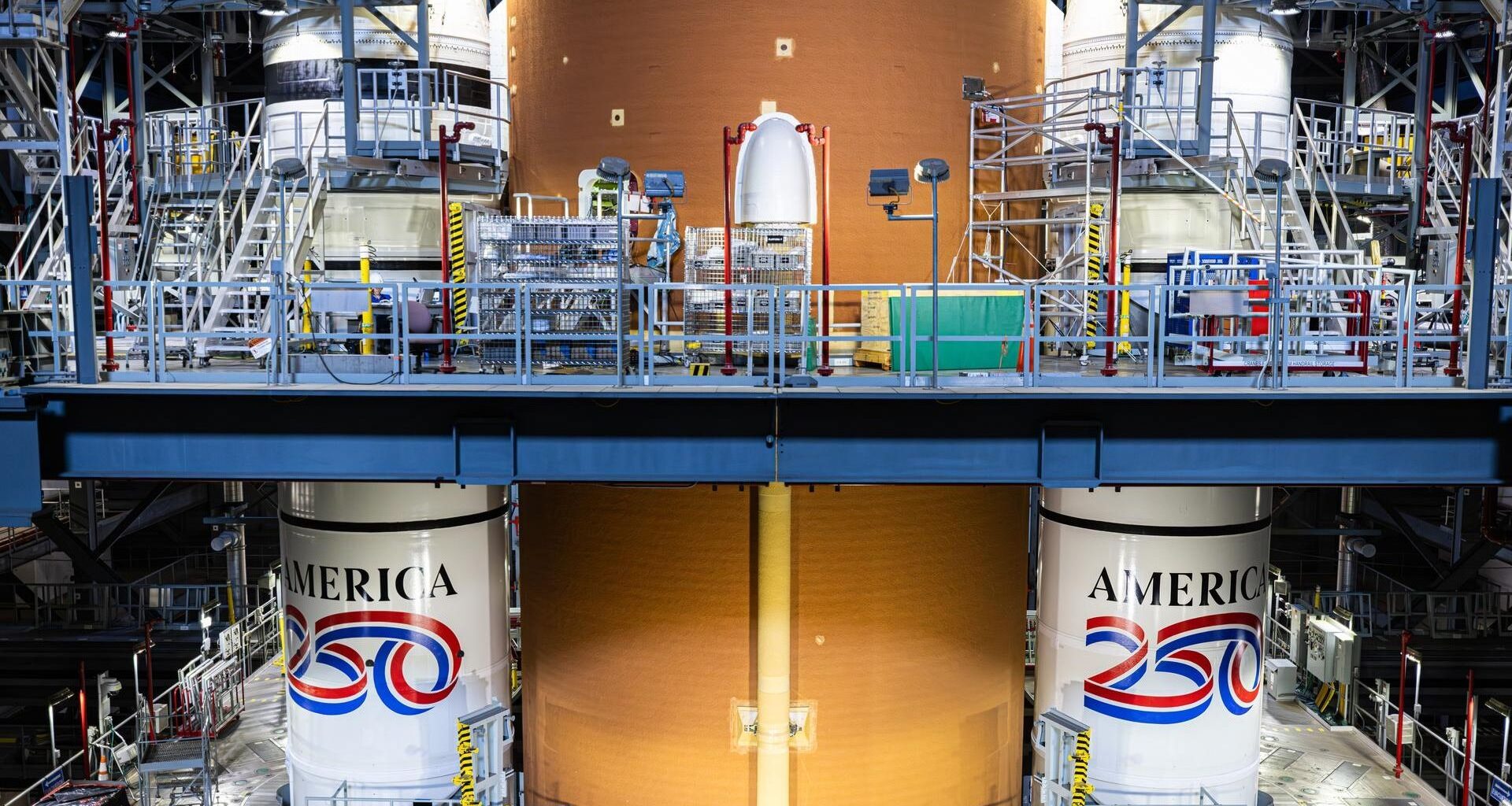 An image from inside a warehouse shows two tall white cylinders on either side of a yellow taller cylinder. The two white cylinders have red, white and blue paint with the words 'America 250' on them.