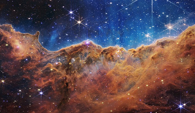 Image showing the orange clouds of the Cosmic Cliffs billowing up into soft peaks in front of a deep blue background. The white sparkle of stars are scattered throughout the image.