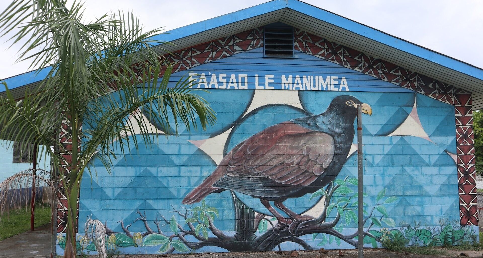Photo of a the exterior of a building on which is a mural of a large bird as viewed from the side. Above the bird it says, "PAASAO LE MANUMEA."