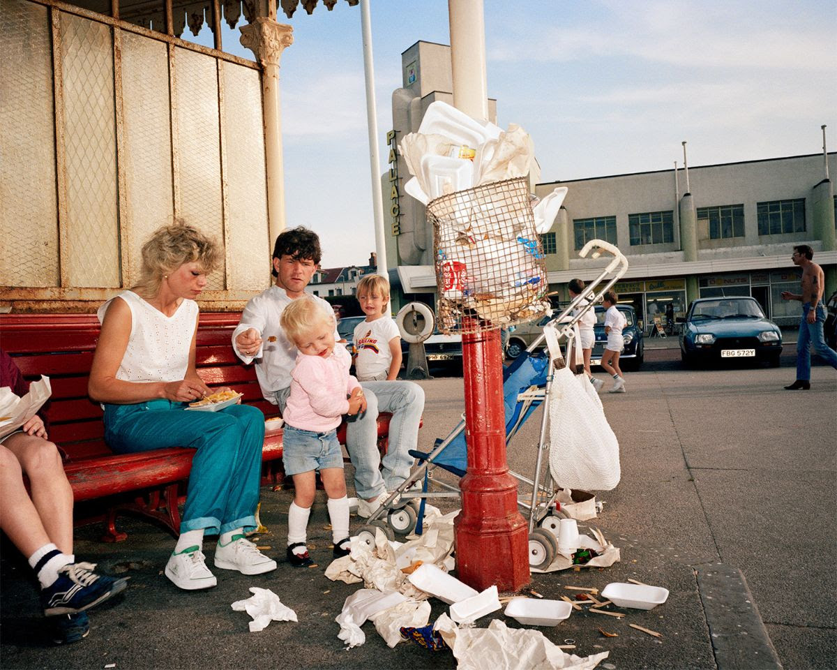 A family sits on a red park bench next to an overflowing red trash can surrounded by discarded food containers and litter.