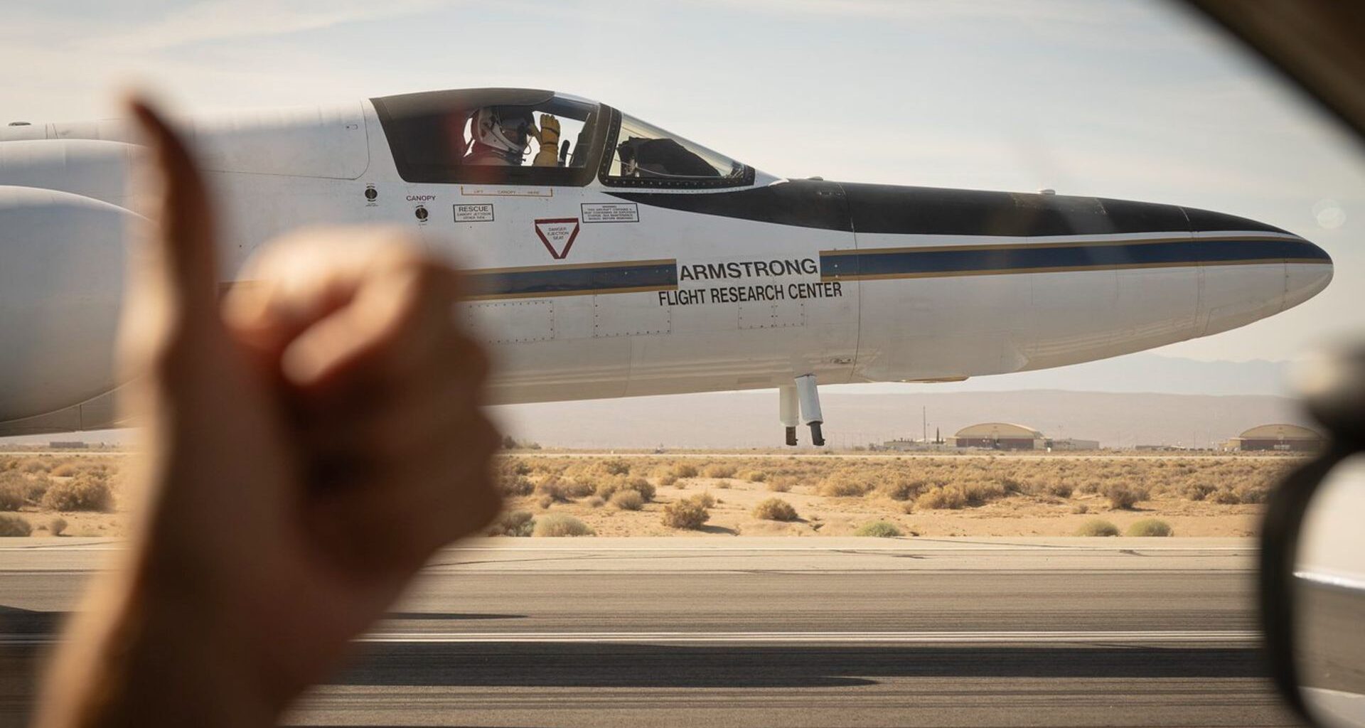 A view from the driver's side window of a car shows a white plane on the runway where the driver's hand shows a thumbs up on the left side of the image.