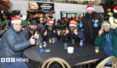A group of homeless people sat around a table, inside a trains station posing with their thumbs up and smiling at the camera