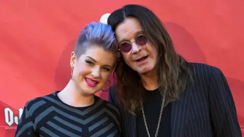 Reuters Kelly Osbourne, with purple hair combed back, wearing a grey and black patterned top, stood on the right of her father in front of a red event backdrop. Ozzy has long black hair, round sunglasses, and is wearing a pinstripe black suit over a black t-shirt.