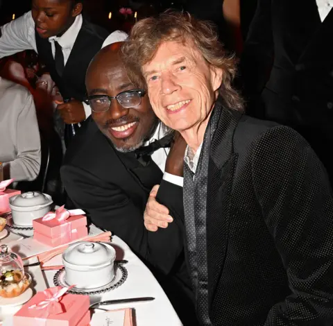 Getty Images Edward Enninful and Mick Jagger sit together at a table in their suits, smiling