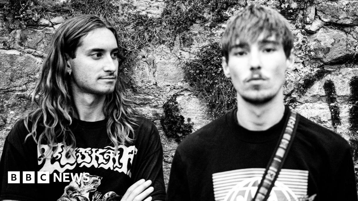 Two young men, one on the left with long hair and one on the right with shorter hair and a light beard, shot in black and white