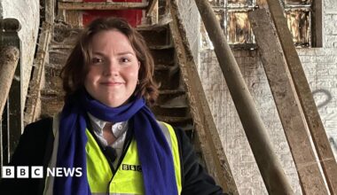 Morgan Ellis Leah stands on a very old wooden staircase. She is wearing a high vis vest and black jacket under it. She has bobbed brown hair and wears a blue scarf.