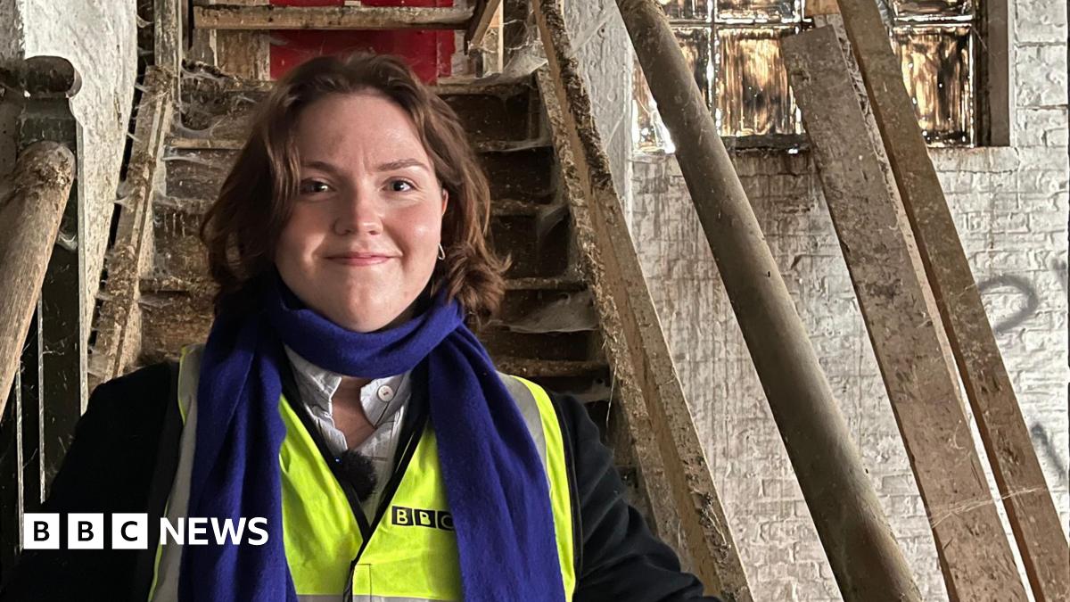 Morgan Ellis Leah stands on a very old wooden staircase. She is wearing a high vis vest and black jacket under it. She has bobbed brown hair and wears a blue scarf.