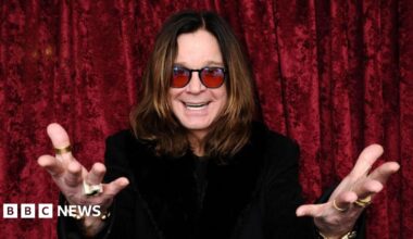 A man with long brown hair and red sunglasses with his hands outstretched and a black top in front of a red curtain
