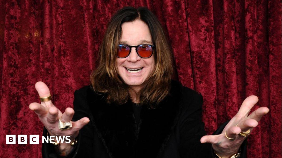 A man with long brown hair and red sunglasses with his hands outstretched and a black top in front of a red curtain