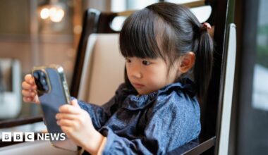 A stock photo of little girl with pigtails looking intently at a smartphone. She is wearing a patterned blue top and sitting in a modern, warmly lit indoor room.