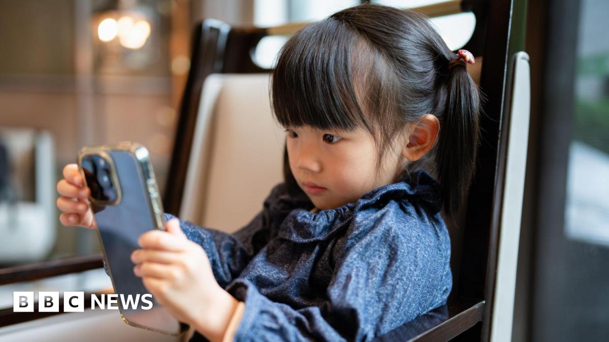 A stock photo of little girl with pigtails looking intently at a smartphone. She is wearing a patterned blue top and sitting in a modern, warmly lit indoor room.