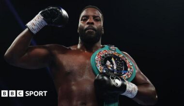 Boxer Lawrence Okolie stands in the ring (which is out of shot) after a fight, posing in a victory stance. Visibly from stomach up, he is bare-chested and muscular. His right arm is bent with his black-gloved fist raised proudly near his head, while his left gloved hand rests on a large championship belt slung over his left shoulder. The belt has a wide teal strap and a shiny, circular centre plate decorated with many small national flags around its edge, catching the arena lights. The background is dark and out of focus, making the boxer and the belt stand out as the clear focal points of the image