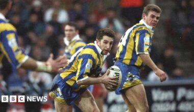 A woman with long light brown hair, dark brown eyes and fringe is smiling into the camera. She has a green, cream and black chequered shirt on. The background is white with nothing else in frame. She has a long blue decoration on her right ear