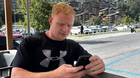 Sol Hyde Sol, wearing a black UnderArmour T-shirt, looks at his phone. He is sitting on a cafe forecourt with a coastal promenade behind him. The weather is sunny.